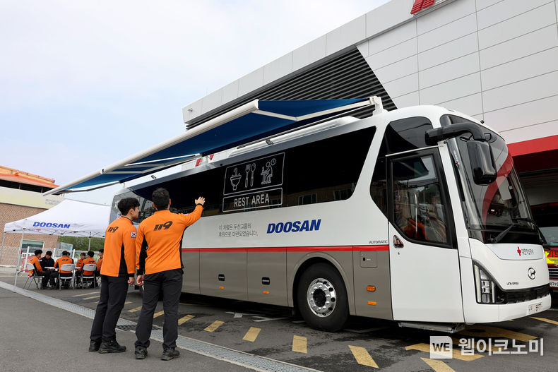 두산 회복버스 외관, 두산이 제작한 ‘재난구호요원 회복버스’를 소방관들이 살펴보고 있다. 차량 내부 뿐 아니라 외부에서도 휴식할 수 있는 전동식 그늘막 장치, 천막과 의자, 테이블이 비치돼 있다. 2023.11.06. (출처: 두산)